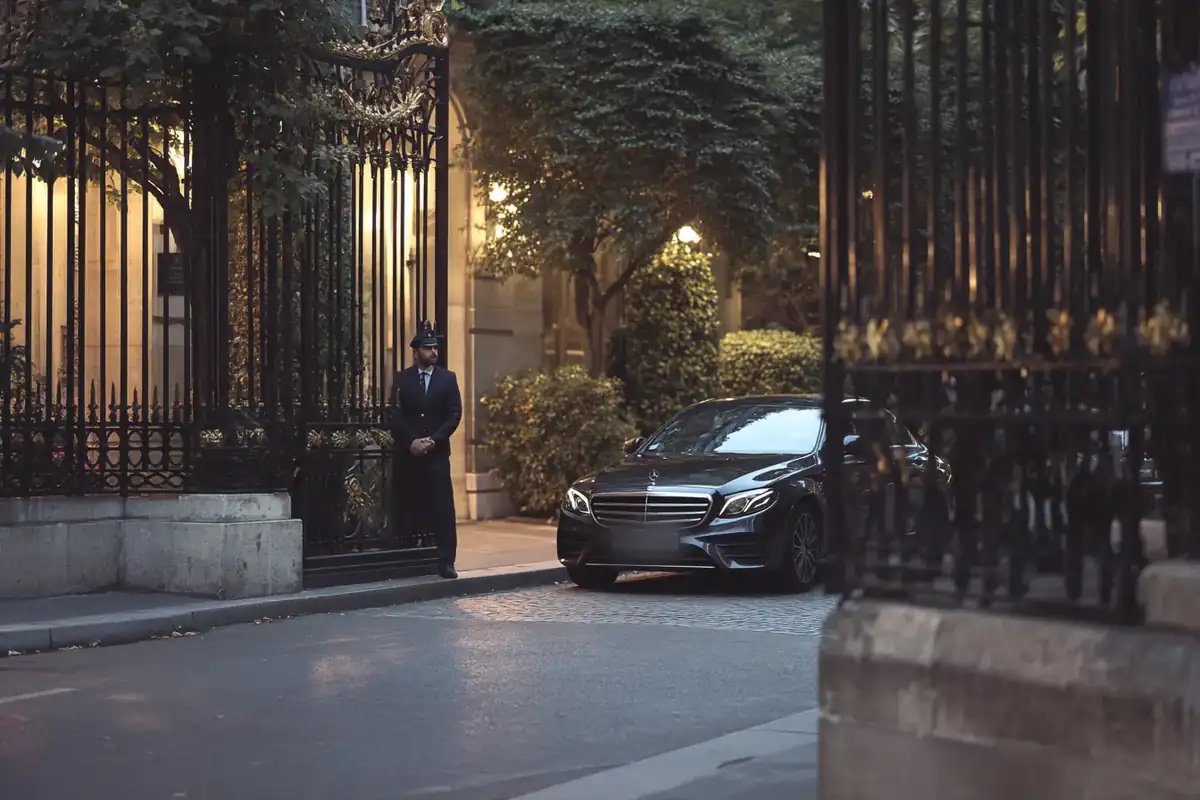 Black sedan beside a wrought-iron garden gate of a Paris luxury hotel in the evening.