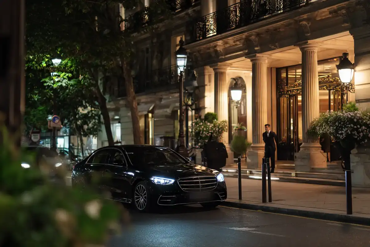 Black sedan arriving at a palace hotel in Paris at night, warm lobby light.