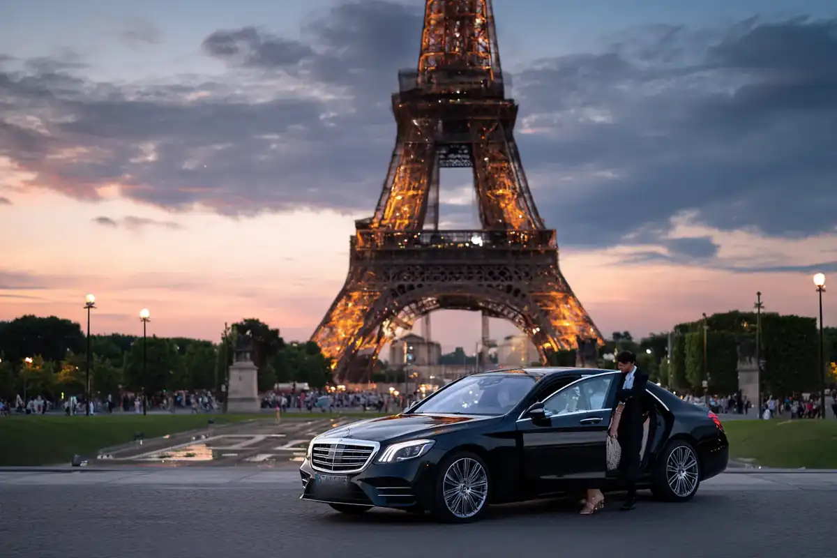 Chauffeur beside a black sedan at the foot of the Eiffel Tower at dusk.