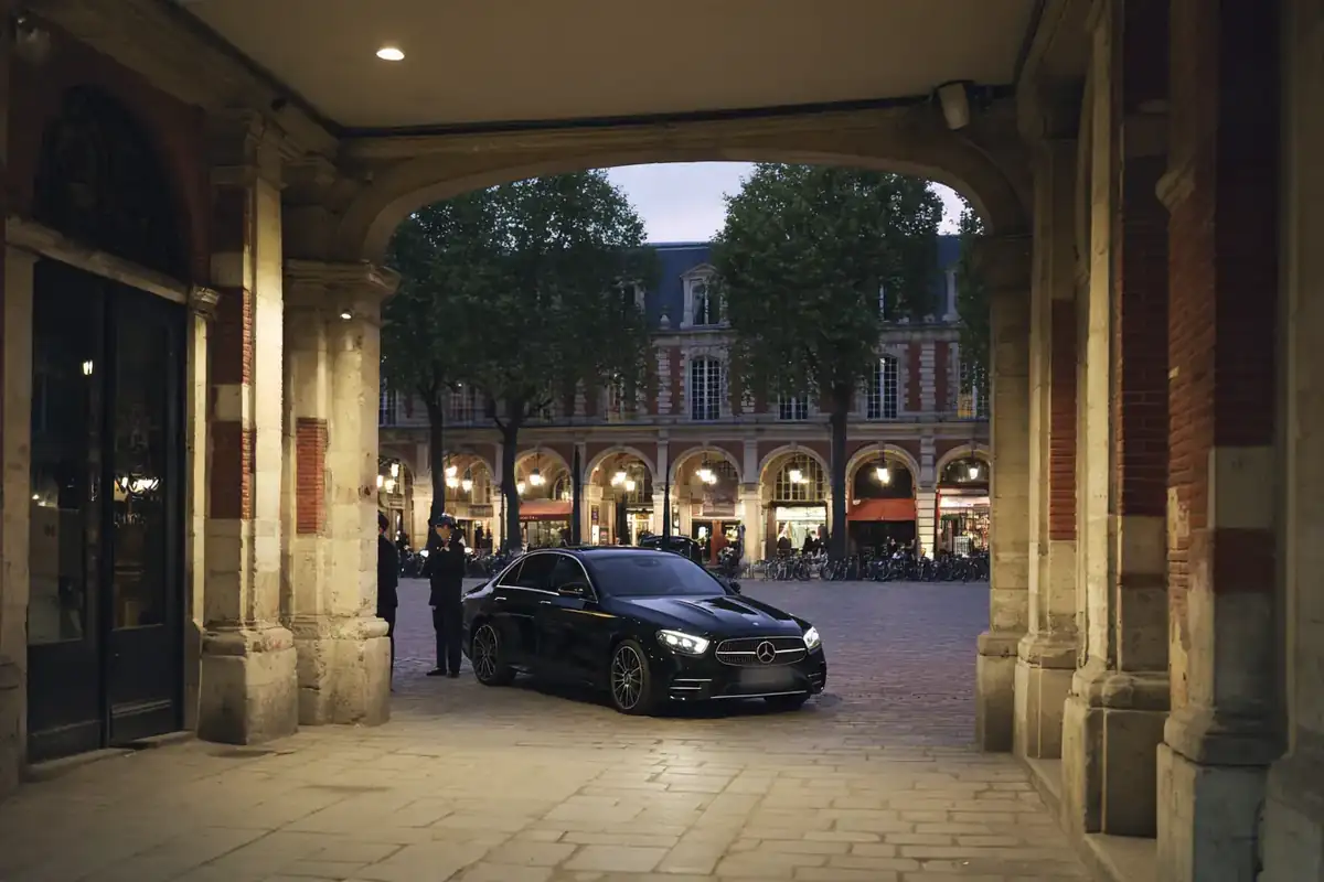 Black sedan before the arcades of a historic Marais square at dusk.