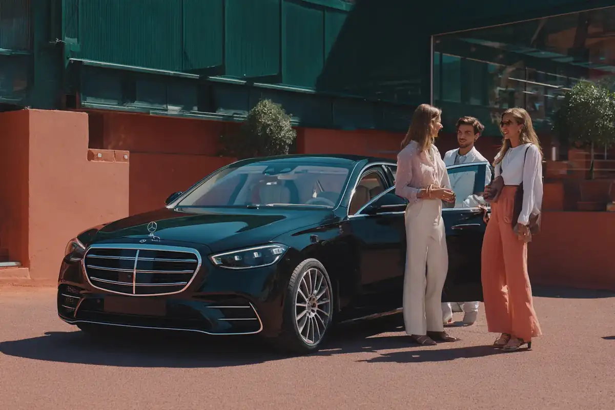 Chauffeur welcoming guests in summer clothes at the open door of a black sedan near a tennis stadium.